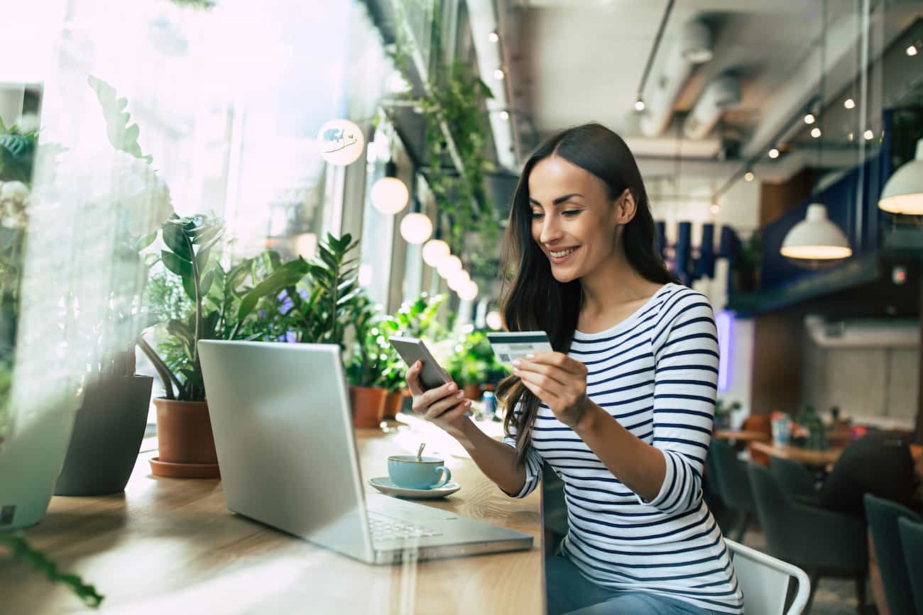 Junge Frau im modernen Café nutzt Smartphone und Laptop, hält eine Kreditkarte, symbolisiert flexible Möglichkeiten, online Geld zu verdienen als Crowdtesterin oder Crowdtester.