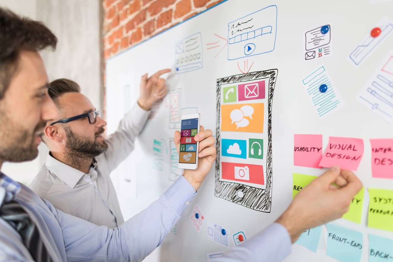 Zwei Männer arbeiten an einem Whiteboard mit farbigen Skizzen und Post-its, um ein digitales Produkt zu testen.