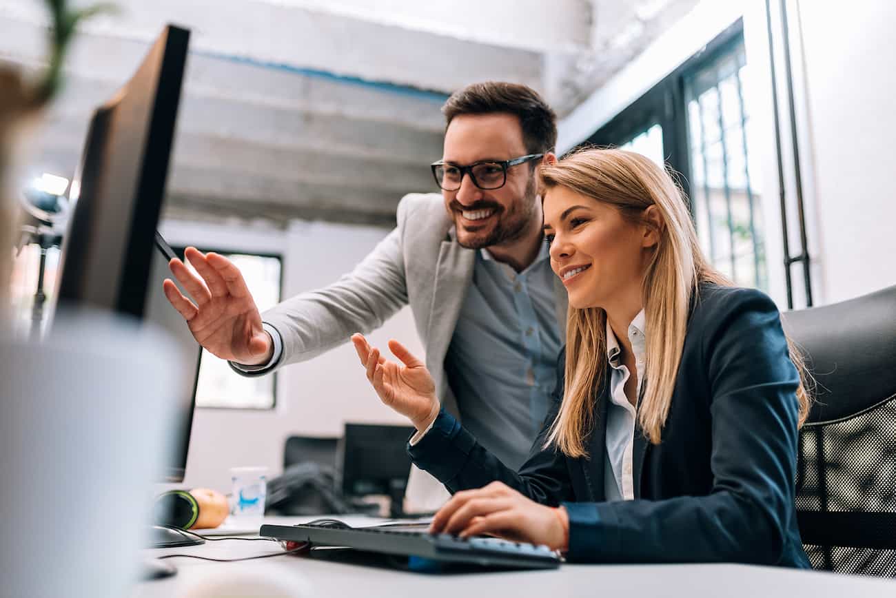 Zwei Kollegen im Büro, die gemeinsam auf einen Computerbildschirm schauen und dabei lächeln, während sie eine Präsentation oder Diskussion über Ergebnisse führen.