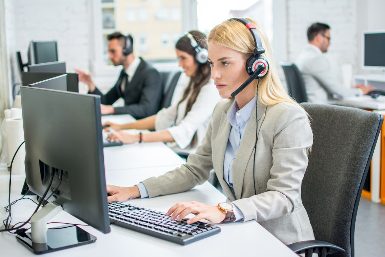 Eine Frau in einem Büro, die ein Headset trägt und konzentriert auf einen Computerbildschirm schaut. Im Hintergrund sind weitere Personen zu sehen, die ebenfalls Headsets tragen und an Computern arbeiten.