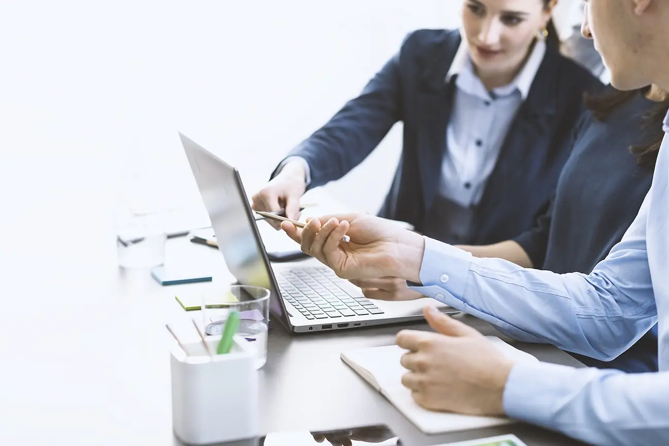 Drei Business-Personen im Meeting am Schreibtisch, eine Person zeigt auf einen Laptop-Bildschirm, während eine andere ein Smartphone hält – symbolisch für koordinierte Softwaretests, Crowdtesting sowie Shift-Left- und Shift-Right-Testing.